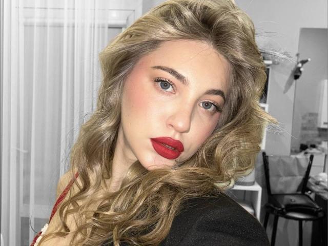 Close-up photo of ShareGullage, a fair-skinned woman with long, wavy blonde hair and bright red lipstick. She's looking directly at the camera with a slightly tilted head, sexual tension evident.