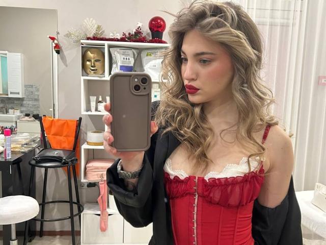 Photographie d'une jeune femme aux cheveux blonds ondulés, au rouge à lèvres rouge et portant un corset en dentelle rouge, tenant un smartphone, en train de prendre une photo. Autoportrait pris devant un miroir dans une pièce très éclairée. Extrêmement sexy..
