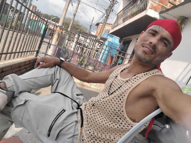 Un uomo latino robusto, muscoloso, con un berretto rosso indossato all'indietro, una canotta di maglia color cuoio, pantaloni grigi, appoggiato a una recinzione di metallo. Sorriso, sfondo urbano illuminato dal sole, fili, edifici. Leggero rigonfiamento nei pantaloni, aspetto casual, sensuale..