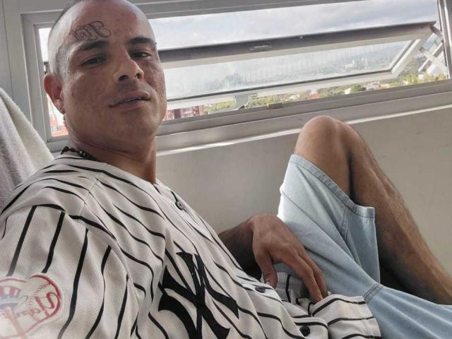 Fotografie eines kahlköpfigen Mannes, StrongAlan, mit einem Tattoo auf der Stirn, der ein gestreiftes Hemd und hellblaue Shorts trägt. Am Fenster sitzend, die Beine gekreuzt, der Penis kaum sichtbar. Lässige, selbstbewusste Pose.