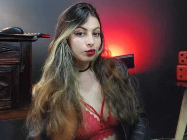 LeilaGoddes, uma mulher curvilínea com longos cabelos castanhos ondulados, usa um sutiã de renda vermelho, batom vermelho e um colar preto. A luz vermelha projeta um brilho sensual em seu rosto. Ela acaricia o decote..