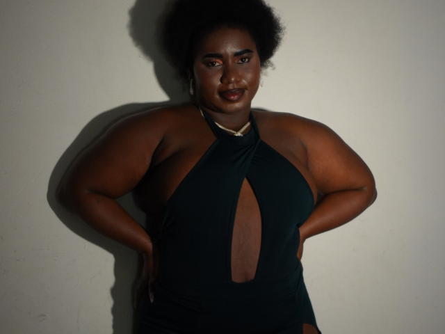 Photograph of a curvy, dark-skinned woman with a large afro, wearing a black halter dress with a large cutout, hands on hips, shadowed against a white wall. Fucking confident.