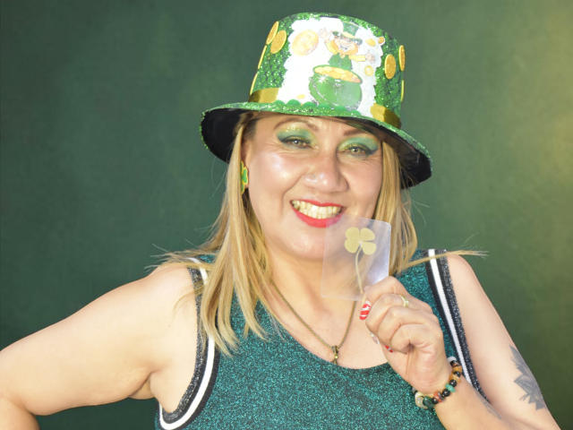 MatureYayita trägt einen grünen, glitzernden Zylinderhut, grünen Lidschatten und ein glitzerndes, ärmelloses Oberteil. Sie hält eine weiße Karte. Eine kleine, gelbe Blume. Lächeln, blondes Haar, Tätowierung am rechten Arm..