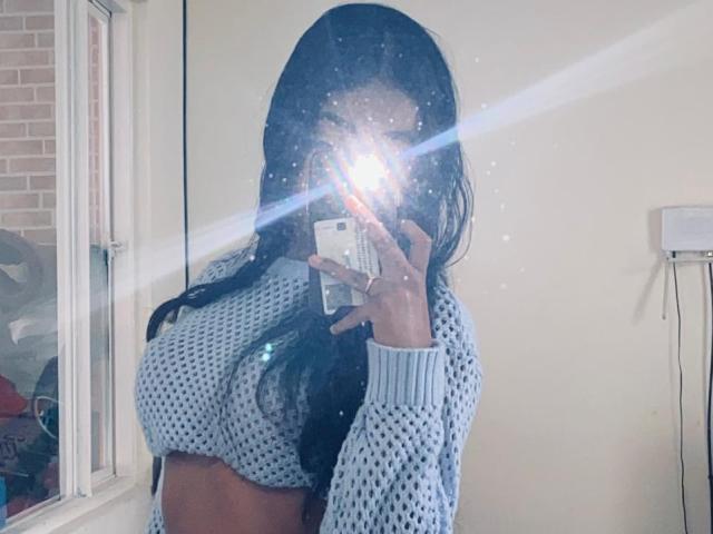 JanneClowses, eine Frau mit langen, schwarzen Haaren, macht ein Selfie mit einem Spiegel, wobei der Blitz ihr Gesicht verdeckt. Sie trägt ein weißes, durchlöchertes Oberteil mit langen Ärmeln, das einen Hauch ihrer Brust zeigt. Helle Lichtblitze.