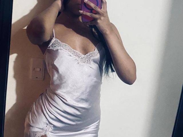 Janne Clowes indossa una sottoveste di seta bianca con bordi in pizzo, e tiene un telefono davanti al viso. La sua mano copre parte del suo petto, suggerendo una posa sensuale..