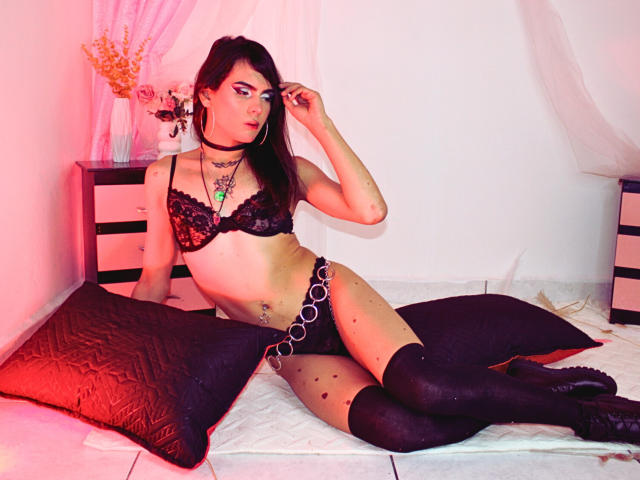 LemlithNyxx, à la peau pâle et aux cheveux noirs, est allongée sur un lit, vêtue d'un soutien-gorge et d'une culotte en dentelle noire, de bas hauts et d'un chaîne de transmission. Des coussins rouges, des rideaux blancs et un vase. Putain, c'est sexy..