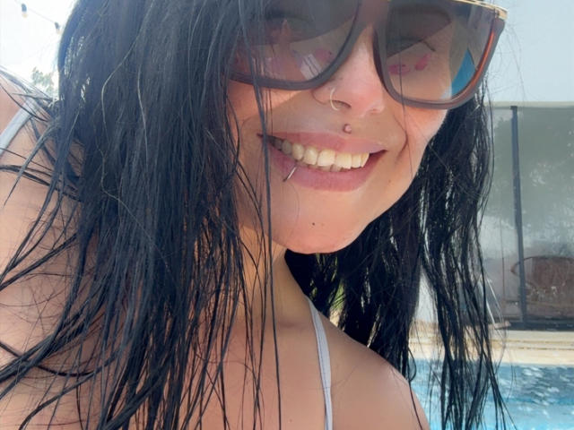 Foto ravvicinata di CharlotteCrofort, con i capelli neri bagnati, che indossa grandi occhiali da sole marroni, un top bianco con spalline, e sorride mostrando il naso. sguardo intenso, luce solare che si riflette sugli occhiali, denti visibili, atmosfera rilassata a bordo piscina..