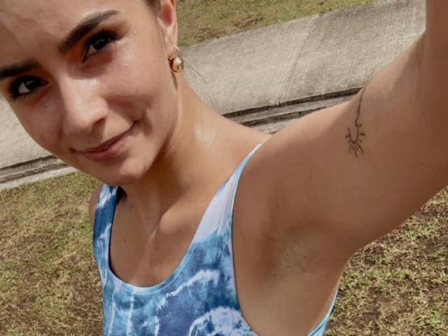 Close-up photo of KimmyK with light brown skin, dark eyebrows, and a small tattoo on her upper arm. She's smiling, wearing a blue tie-dye tank top, outdoors near a sidewalk.