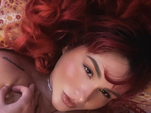 Close-up of DelirioPerpetuo lying on a patterned surface, head tilted, red curly hair, intense gaze, subtle makeup, hand near shoulder, tattoo on upper arm, sultry expression, slight blush.