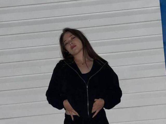 MikeBooras, der vor einer weißen Wand mit horizontalen Streifen steht, trägt eine schwarze Jacke mit Reißverschluss, die Hände sind an den Hüften, er hat langes, dunkles Haar. Haare, halb geschlossene Augen, leicht nach hinten geneigt, vermittelt eine lässige Selbstsicherheit und subtile sexuelle Spannung..