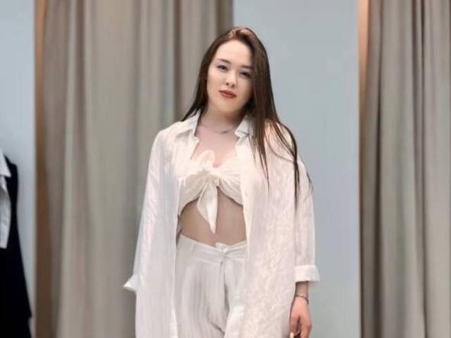 Mike Booras, con cabello negro largo, vistiendo una camisa blanca abierta que revela una camiseta blanca anudada y pantalones blancos a juego. De pie, con una expresión segura, una ligera sonrisa, y una iluminación suave..