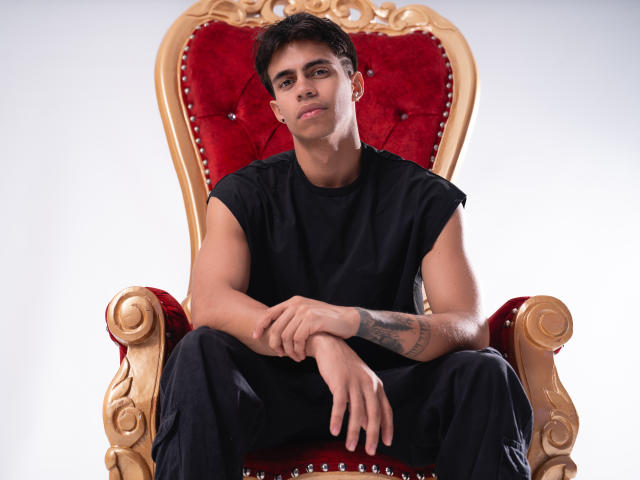 CodyPipper, muscular and sexy, sits on an ornate, red velvet throne with gold trim. He wears a black t-shirt, arms tattooed, legs spread, cock partially visible, intense gaze.
