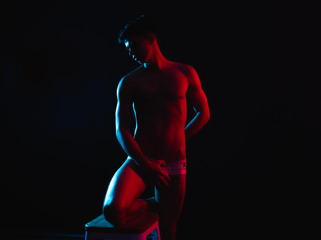 CodyPipper, shirtless, muscular, standing in shadowy room, illuminated by blue and red lights, leaning on chair, wearing only tight, red briefs, hand resting on crotch, intense, sexual tension.