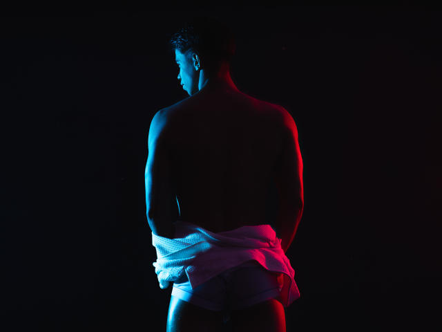 CodyPipper, muscular male, backlit with blue and red lights, wrapped in a white cloth, standing with hands behind back, shadowed face, intense lighting, stark contrast, sexual tension, dark background.