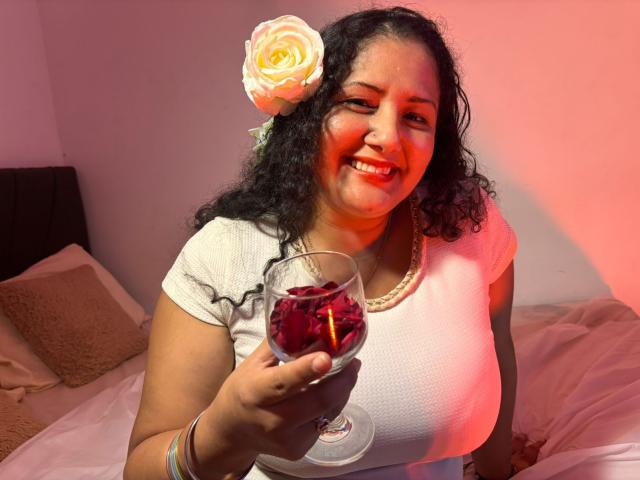 GenesisMontenegro, a curvy woman with dark curly hair and a white rose in her hair, smiles while holding a glass of red wine. She's wearing a white top, glowing under red light.