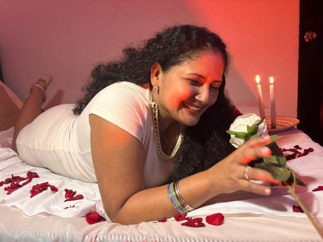 GenesisMontenegro, een mollige, donkerhuidige vrouw met lang zwart haar, liggend op een wit bed, een witte roos vasthoudend, omringd door... door rode rozenblaadjes, rood licht op de achtergrond, lachend, aantrekkelijk, kaarsen in de buurt..