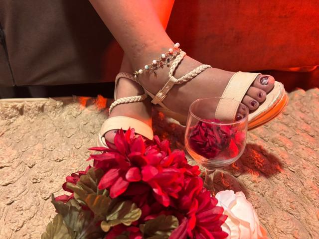 Les jambes de GenesisMontenegro, chaussées de sandales blanches à fines lanières ornées de strass, les pieds croisés, avec des ongles peints en violet, et un verre à la main. Pétales de rose rouge, fleurs rouges, surface beige texturée, lumière rouge, ambiance sensuelle, érotique..