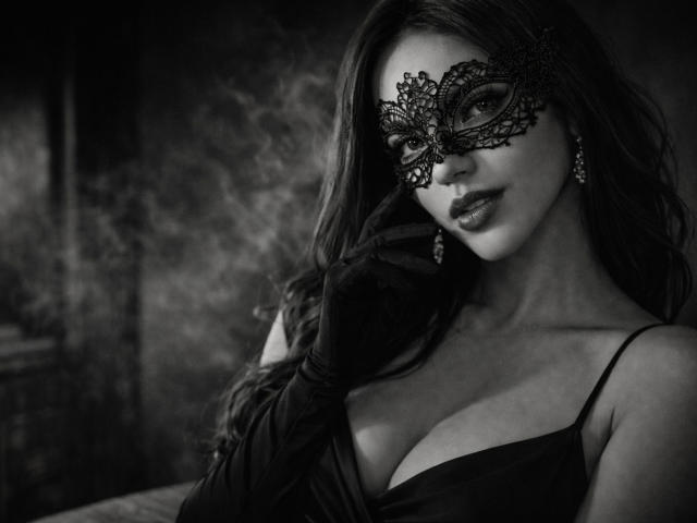 Black and white photograph of SofieWhite with long hair, wearing a lacy black masquerade mask, dark lipstick, and a low-cut black dress, showing cleavage. Smoky background, sensual pose.
