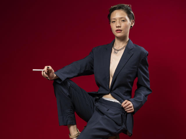 A LissyLuxe está posicionada contra um fundo vermelho escuro, vestindo um terno azul escuro com um cinto branco, sem camisa, expondo sua fenda. Ela segura um cigarro na mão direita, com as pernas cruzadas, transmitindo confiança e sensualidade..