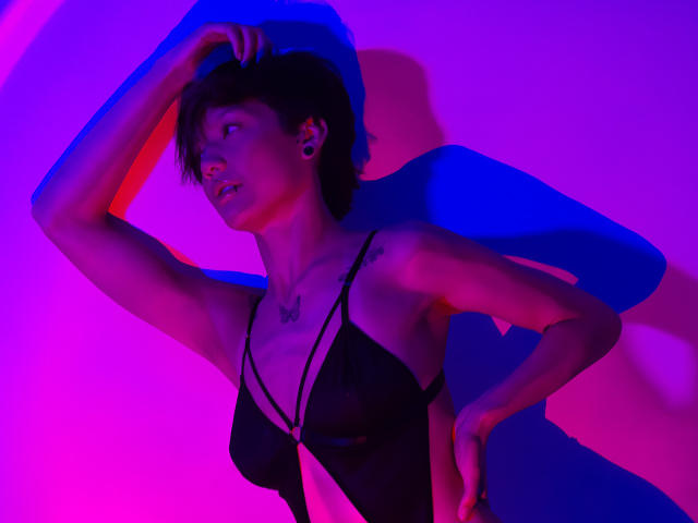 LissyLuxe, mit kurzen, schwarzen Haaren, trägt ein schwarzes, durchsichtiges Oberteil und posiert mit einem hochgehobenen Arm, im Hintergrund leuchten blaue und pinke Neonlichter. Ihre Figur betonen. Schattenspiele verleihen Tiefe und eine intensive, erotische Atmosphäre..