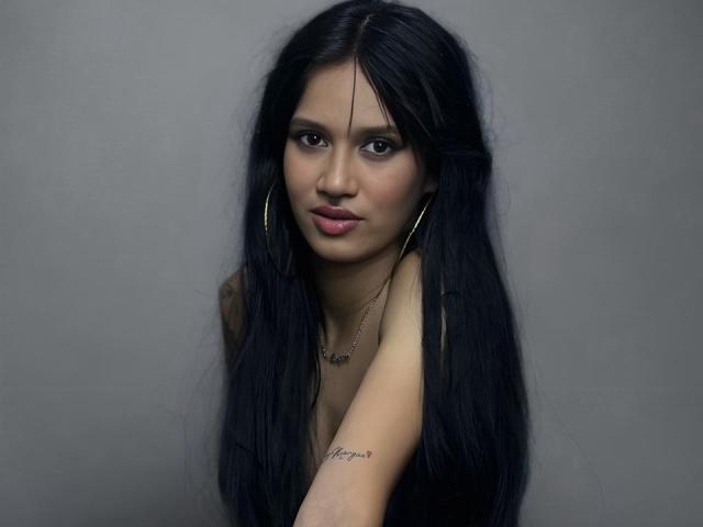 LucyMoorgana hat langes, schwarzes Haar, durchdringende Augen und rosafarbene Lippen. Sie trägt große Ohrringe im Stecker-Stil und eine Halskette. Sie hat ein Tattoo an ihrem rechten Arm. Grauer Hintergrund. Verdammt sexy, blickt direkt in die Kamera..