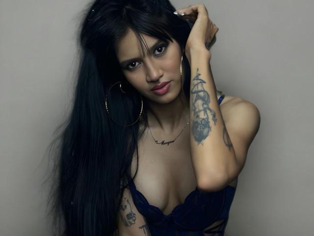 LucyMoorgana, dunkelhaarig, langes Haar, stechende Augen, schwarzes Spitzenoberteil, kleine Brüste, Tattoos am linken Arm, Hand im Haar, silberne Hängeohrringe, dezentes Make-up, grauer Hintergrund, sexy, verführerische Pose, selbstbewusster Ausdruck..