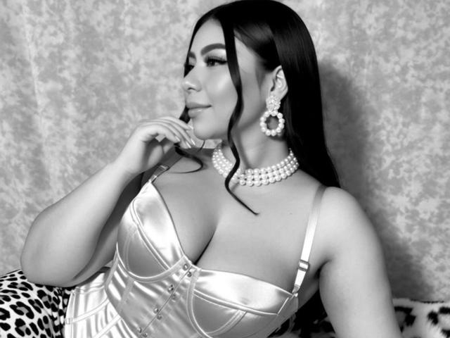 Foto en blanco y negro de Gabriela Ferrer con cabello negro largo, vistiendo un vestido brillante y escotado, aretes grandes y collar ajustado al cuello. Ella está apoyando el mentón en la mano, luciendo seductora. El fondo tiene un patrón texturizado..