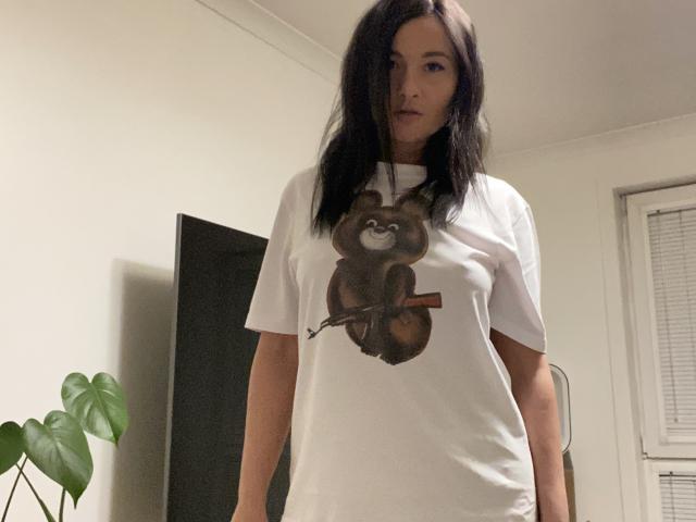 Dionisia with black hair, wearing a white t-shirt with a sloth graphic, stands in a room with a potted plant and window blinds. She looks directly at the camera, exuding sexual tension.