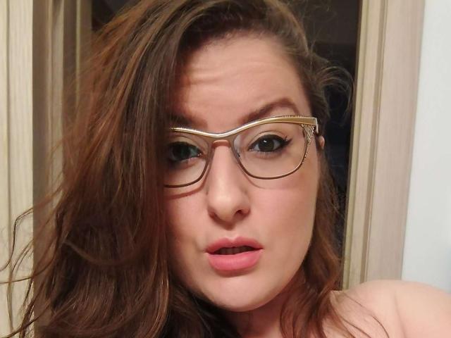 Close-up of BadAngel, a woman with brown hair, wearing gold-rimmed glasses, looking directly at the camera with slightly parted pink lips. Her expression is suggestive and slightly intense.