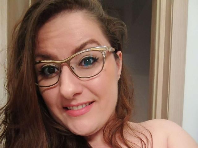 Close-up van BadAngel met lang bruin haar, blauwe ogen en een bril met gouden rand, die lacht met roze lippenstift. De blik van BadAngel is direct, met een lichte blos op de wangen, wat subtiele seksuele spanning suggereert..