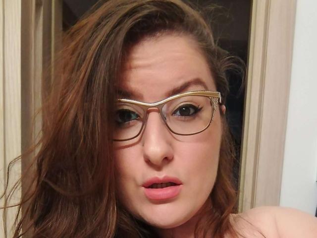 Primer plano de BadAngel, una mujer de piel clara y cabello castaño, que lleva gafas con montura dorada, labios ligeramente entreabiertos, y expresión de sorpresa. Enfoque en el rostro, fondo desenfocado. Increíblemente sexy..