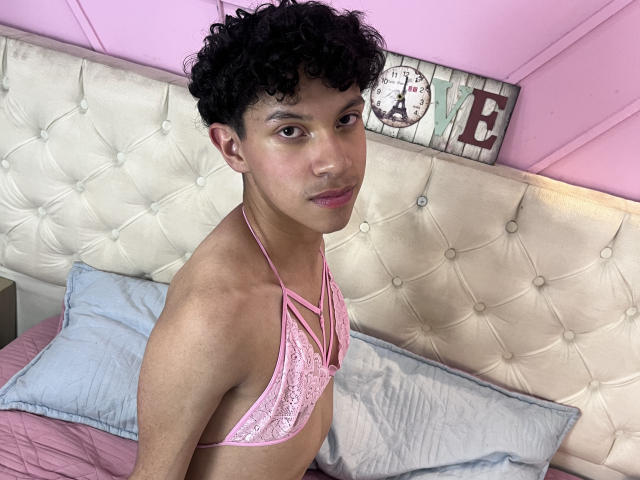 Fotografia de um jovem, corpulento, com cabelo curto e encaracolado, de cor preta, vestindo um sutiã de renda rosa, sentado em uma cama com um edredom. cabeceira. A luz rosa emite um brilho sedutor..