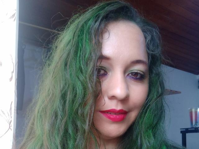 Photo en gros plan de NalimiSmirnov, avec des cheveux verts et ondulés, du rouge à lèvres rouge et du fard à paupières violet. Son visage est légèrement incliné, révélant un sourire subtil. La pièce en arrière-plan est faiblement éclairée. Tension sexuelle due au regard direct.