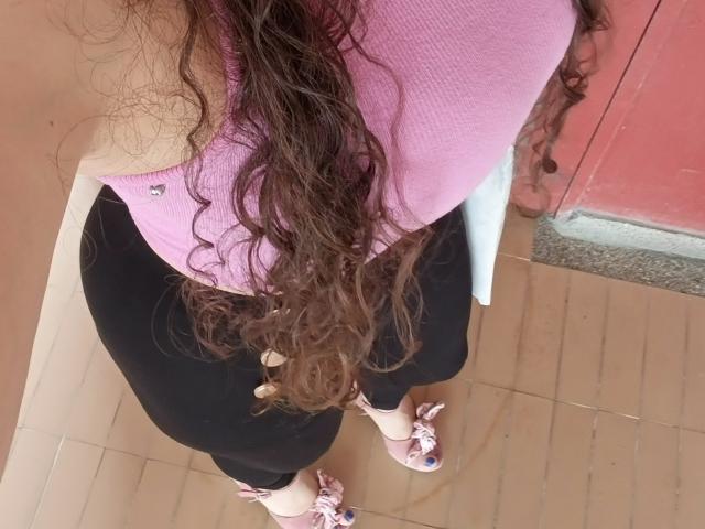 Visão de cima de NalimiSmirnov, mostrando cabelos castanhos longos e ondulados, uma blusa rosa, uma saia preta e tênis rosa. Concentre-se na curva de suas nádegas e na tensão em sua postura..