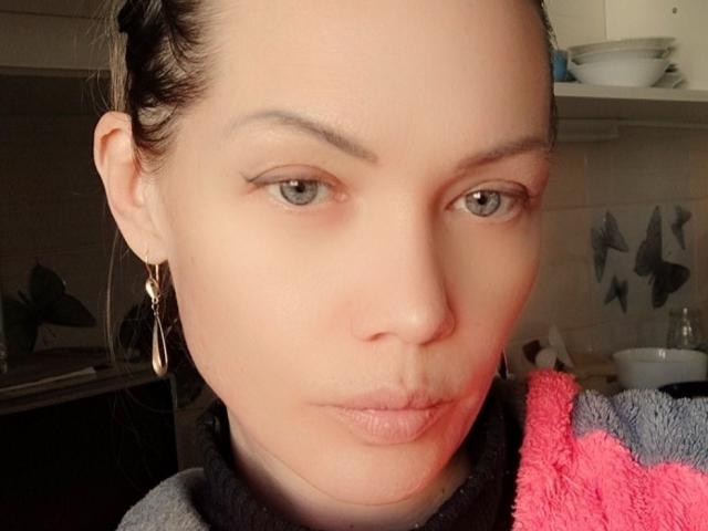Close-up of JuliaLoves with pale skin, brown eyes, and subtle makeup. She has small, slightly puckered lips and wears gold earrings. Her dark hair is pulled back. A red and gray towel is visible.