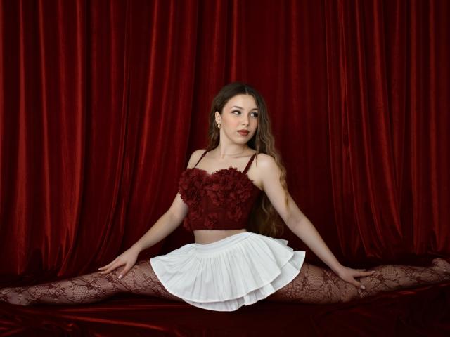 LilyBlake, seduta con le gambe divaricate su un divano di velluto rosso, indossa un top corto floreale di colore rosso scuro e una gonna bianca plissettata. I suoi lunghi capelli castani le ricadono sulle spalle. Si percepisce una forte tensione sessuale..