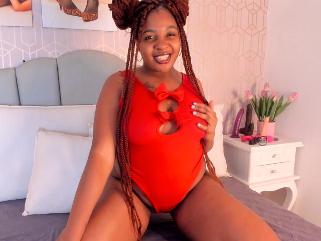 Xhaniyah, uma mulher negra de corpo curvilíneo com longas tranças, está sentada em uma cama vestindo um biquíni vermelho com recortes, sorrindo de forma sedutora. Suas pernas estão abertas, revelando suas coxas. Uma mesinha de cabeceira branca com flores está ao fundo..