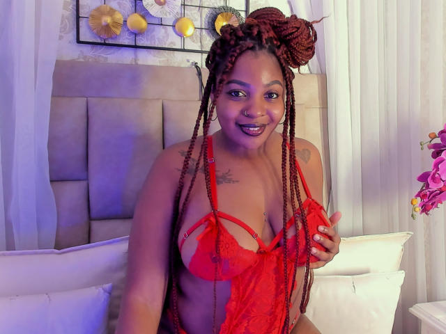 Photographie de Xhaniyah, à la peau foncée, avec de longs cheveux tressés, portant un haut en dentelle rouge qui expose sa petite poitrine. et décolleté. Elle sourit, assise sur un lit avec des oreillers blancs..