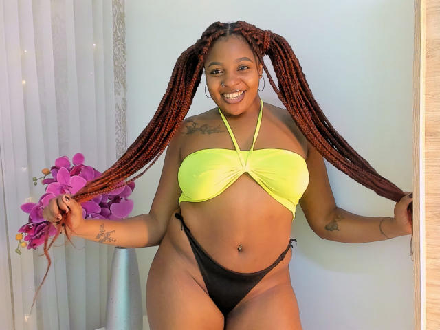 Xhaniyah está de pie con confianza, con su larga trenza, sujetándola por ambos lados. Ella lleva un top de bikini amarillo y una tanga negra, y está sonriendo. El fondo incluye flores moradas y cortinas blancas. Iluminación brillante.