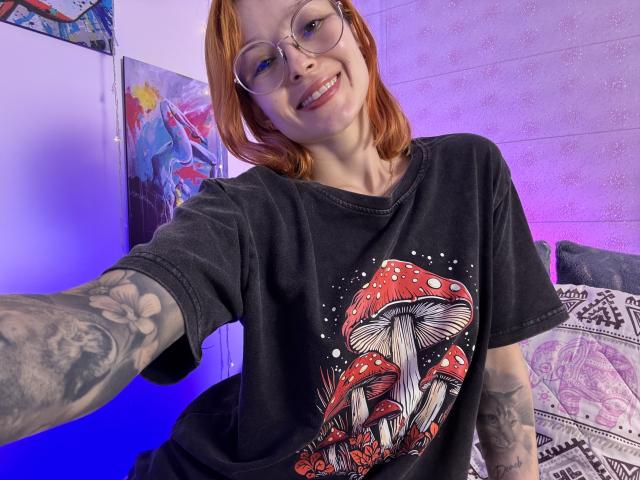 Sarah Rousen, con cabello rojo y gafas, sonriendo, vestida con una camiseta negra con un gráfico de un hongo rojo, con un brazo tatuado. Inclinándose hacia adelante, con el pene erecto visible en unos pantalones cortos negros, iluminación púrpura y azul..