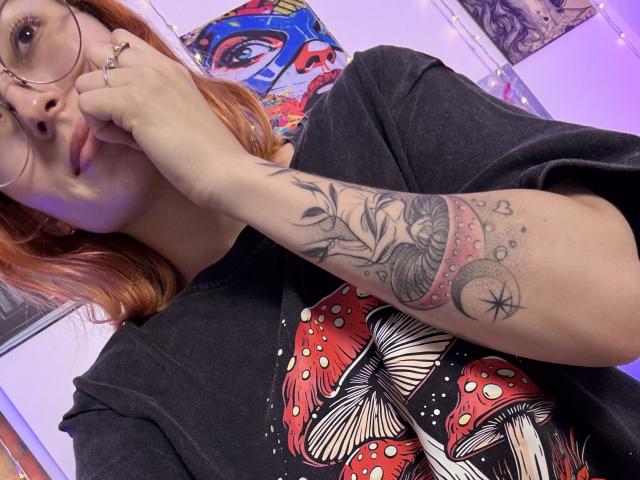 Sarah Rousen, Brille, rötlich-braunes Haar, schwarzes T-Shirt mit rotem und weißem Aufdruck, tätowierter Arm mit aufwendigem Design, hand in der Nähe des Gesichts, leicht gerötet, lässige Pose, lebendige Farben, moderner Stil..