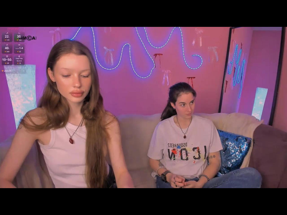 Two women sitting on a couch in a brightly lit room. KhloeToDaisy on the left, long brown hair, white tank top, relaxed pose. Other KhloeToDaisy, ponytail, "Texas State" shirt, looking at camera. Neon lights in background.