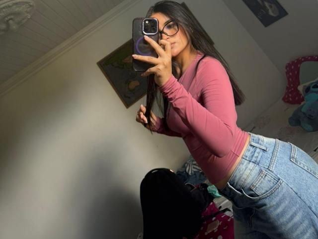 MiiaPalmer, une femme aux formes généreuses avec de longs cheveux noirs et des lunettes, prend un selfie devant un miroir. Elle porte un haut rose moulant à manches longues et un jean taille haute, ce qui met en valeur ses fesses volumineuses. Extrêmement séduisant..