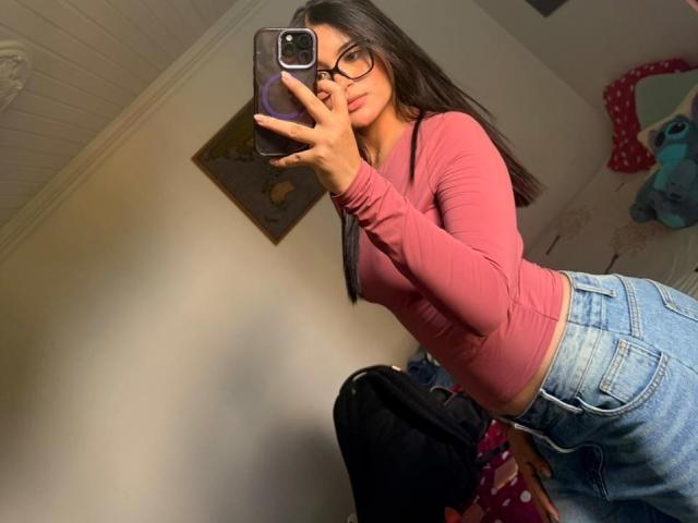 MiiaPalmer se toma un selfie en una habitación con poca luz, vistiendo una camiseta rosa ajustada de manga larga y unos vaqueros azules de cintura alta. Ella tiene el pelo largo y negro, usa gafas y sostiene un teléfono inteligente. Arruina la composición..