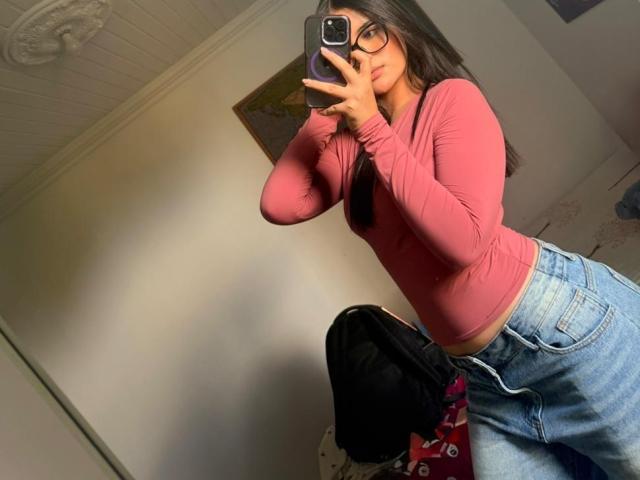 MiiaPalmer, une femme séduisante avec de longs cheveux noirs, portant un haut rouge moulant à manches longues et un jean bleu taille haute, prend autoportrait pris dans un miroir. Sa main est sur son pénis, le téléphone dans l'autre..