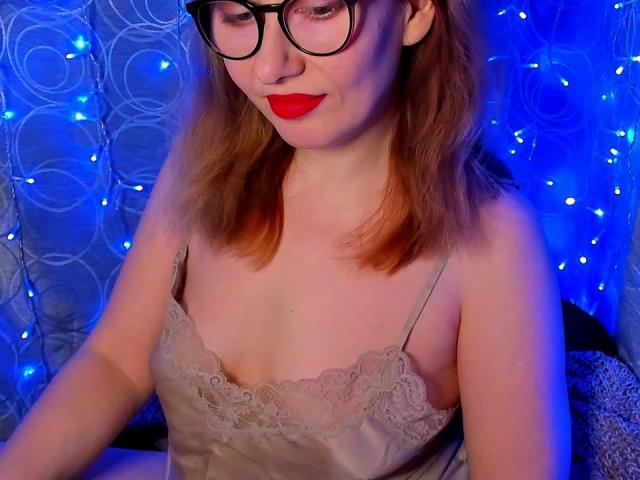 PaulinaSunny, fair-skinned with red lipstick, wearing black-rimmed glasses and a lace-trimmed beige camisole, looks down with a slight smirk. Blue fairy lights and patterned backdrop create a sensual atmosphere.