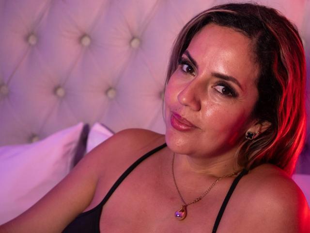 Close-up of LorenaSoto, a curvy woman with tan skin and dark brown hair, wearing a black tank top, glistening with sweat, red and purple lighting, tufted headboard, seductive smile.