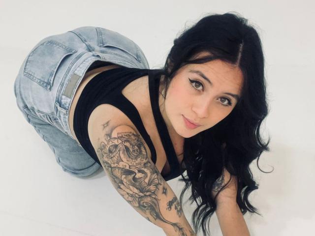 AnnyBentleii, eine Frau mit langen schwarzen Haaren, einem tätowierten Arm und einem schwarzen Oberteil, befindet sich auf allen Vieren und blickt verführerisch nach oben. Blick. Sie trägt Jeansshorts mit hohem Bund, die ihre Figur betonen..
