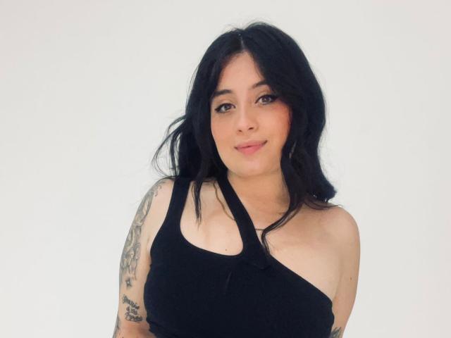 Fotografía de Anny Bentleii con cabello negro hasta los hombros, piel clara y tatuajes en su brazo derecho. Ella lleva una blusa negra con un solo hombro, sonriendo ligeramente, con un evidente indicio de tensión sexual..