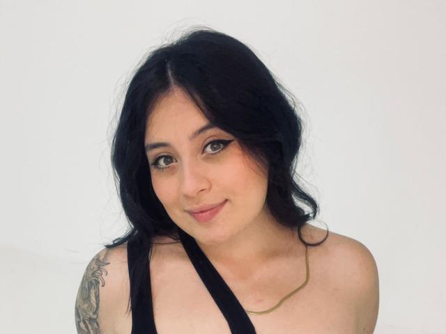 Fotografia de Anny Bentleii, uma jovem com cabelos longos e pretos, pele clara e uma tatuagem no ombro direito. Ela está sorrindo levemente, vestindo uma blusa preta com alças e uma corrente dourada. Muito fofo.